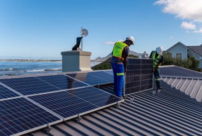 Two African men install solar panels neatly on a residential roof of a house near the ocean. Sustainable living.